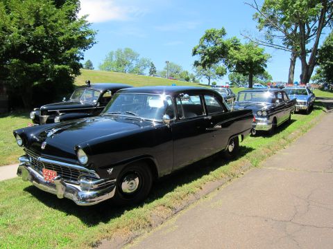 1991 Ford LTD Crown Victoria - black mid-1950s Ford State Police sedan with red plate parked on grass beside a curb, other cruisers behind.