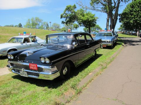 1991 Ford LTD Crown Victoria - black late-1950s Ford State Police sedan with red front plate parked on grass beside a paved drive.