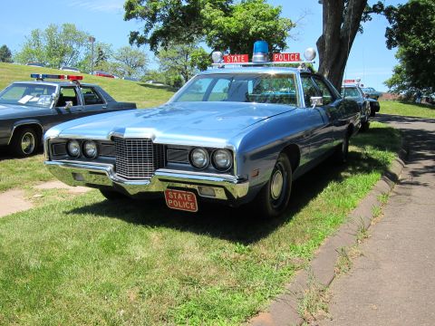 1991 Ford LTD Crown Victoria - light-blue 1970s Ford State Police sedan with roof light bar parked on a grassy field at a car show.