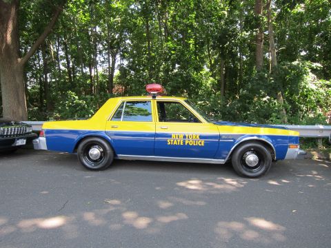1991 Ford LTD Crown Victoria - blue-and-yellow New York State Police sedan with roof light shown in side profile on a paved road by trees.