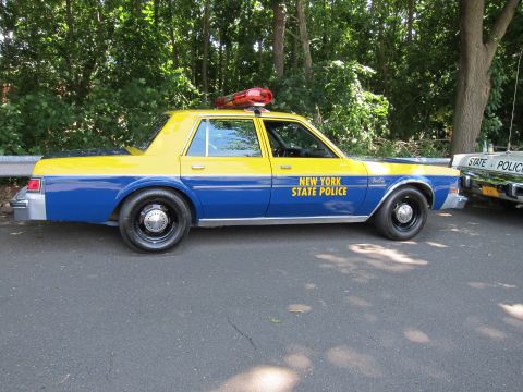 1991 Ford LTD Crown Victoria - blue-and-yellow New York State Police sedan with a red roof light parked on pavement beside a guardrail and trees.