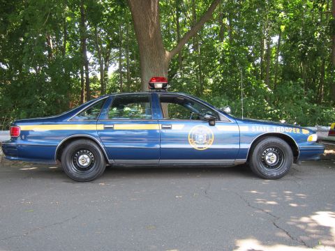 1991 Ford LTD Crown Victoria - dark-blue Chevrolet Caprice State Trooper sedan with gold door seal and roof light parked on pavement near trees.