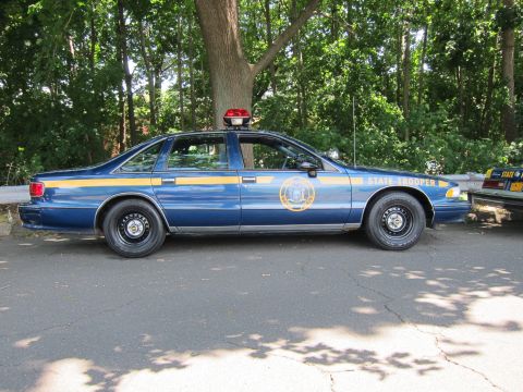 1991 Ford LTD Crown Victoria - dark-blue Chevrolet Caprice State Trooper sedan with gold seal parked on a road beside a guardrail and a yellow cruiser.