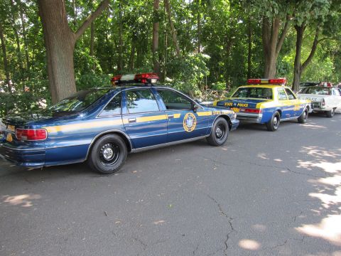 1991 Ford LTD Crown Victoria - blue Chevrolet Caprice State Trooper sedan parked behind yellow-and-blue State Police cruisers on a shaded road.