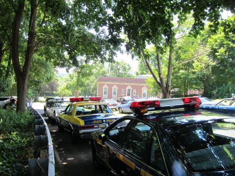1991 Ford LTD Crown Victoria - line of vintage police cruisers with lightbars parked along a tree-shaded drive in front of a low brick building at a car show.