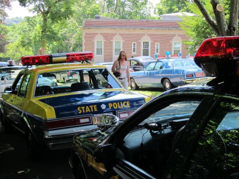1991 Ford LTD Crown Victoria - yellow and blue STATE POLICE sedan parked beside a black police car, with a woman standing between vehicles at an outdoor show.