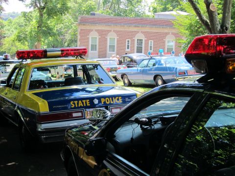 1991 Ford LTD Crown Victoria - yellow and blue STATE POLICE sedan beside a black cruiser, with classic cars and a brick building visible in the background.