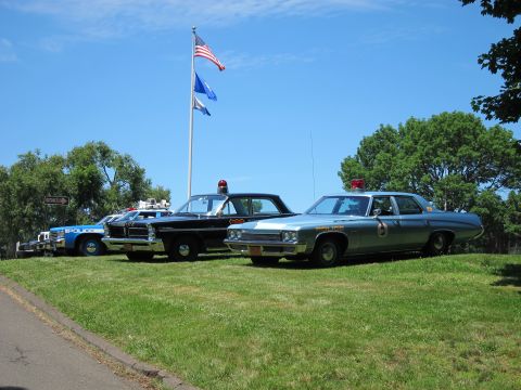 1991 Ford LTD Crown Victoria - Three vintage police cars, including a black-and-white and a silver cruiser, parked on grass beneath a US flag and a state flag.