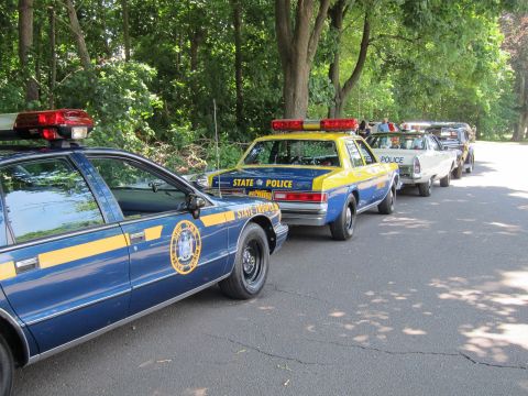 1991 Ford LTD Crown Victoria - line of police cruisers, including blue STATE POLICE cars and a Crown Victoria, parked along a tree-lined paved road.
