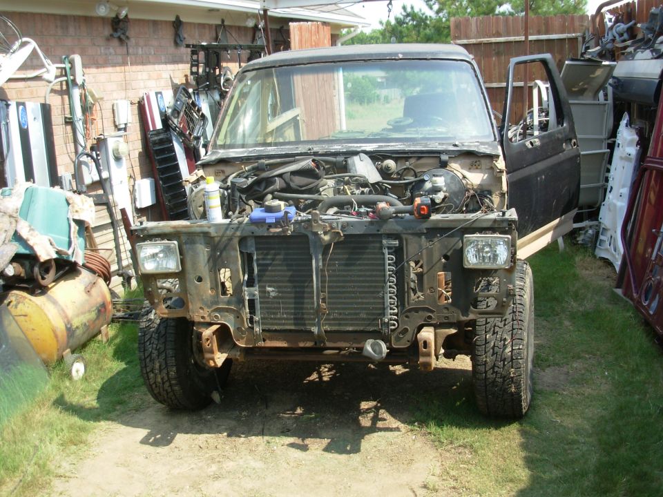 Front of dark Ford Bronco with front end stripped, radiator and engine exposed, in a junk yard.