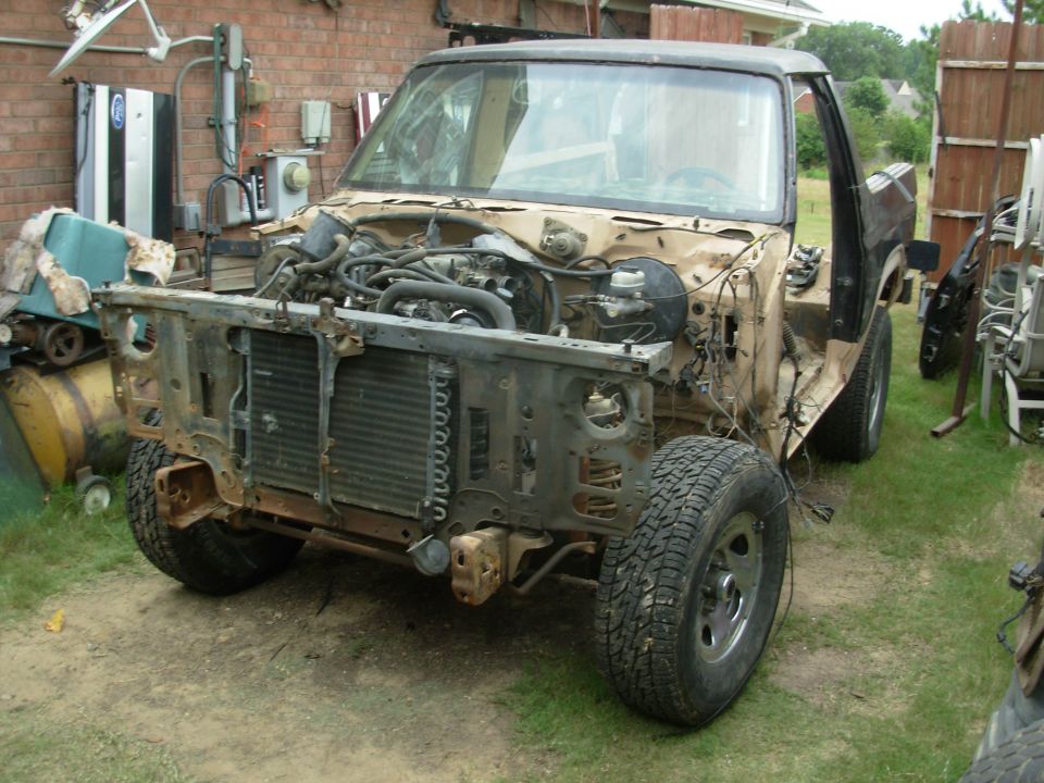 Partially disassembled tan Ford truck with front end stripped, parked in a backyard.