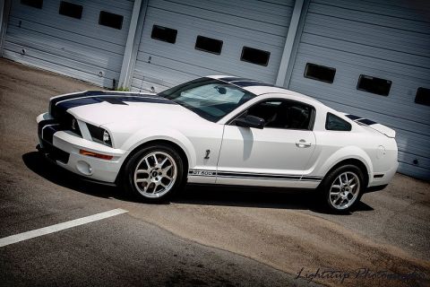 2007 Ford Mustang - A white 2007 Ford Mustang GT 500 with black racing stripes is parked in front of a large garage door.