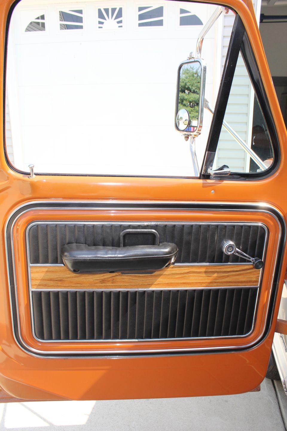 Orange truck door interior with black ribbed upholstery, wood trim, and chrome handle. Visible side mirror reflects trees