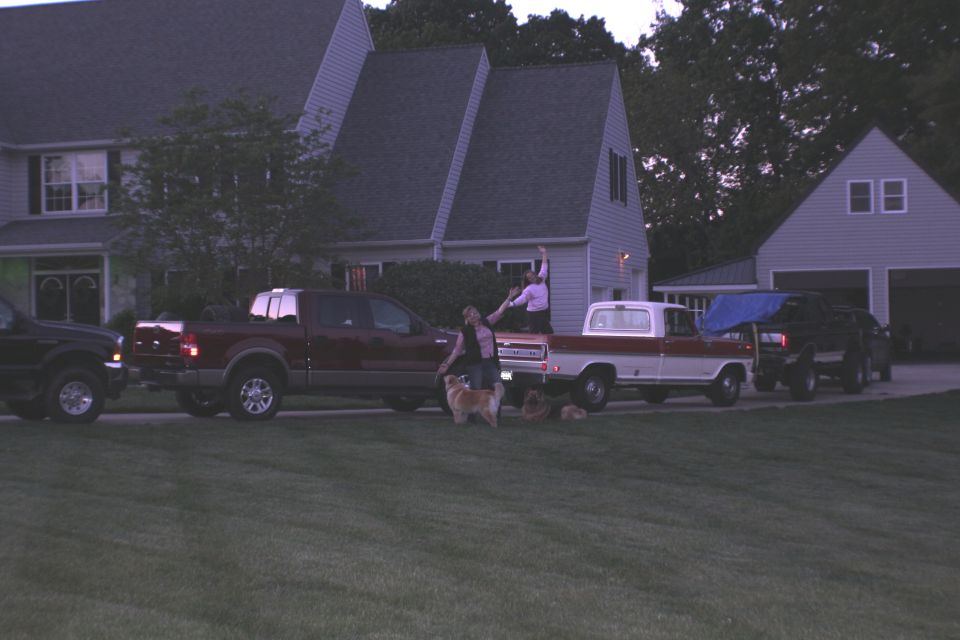 Dusk driveway with black SUV, red crew-cab pickup, vintage red and white Ford pickup and dogs