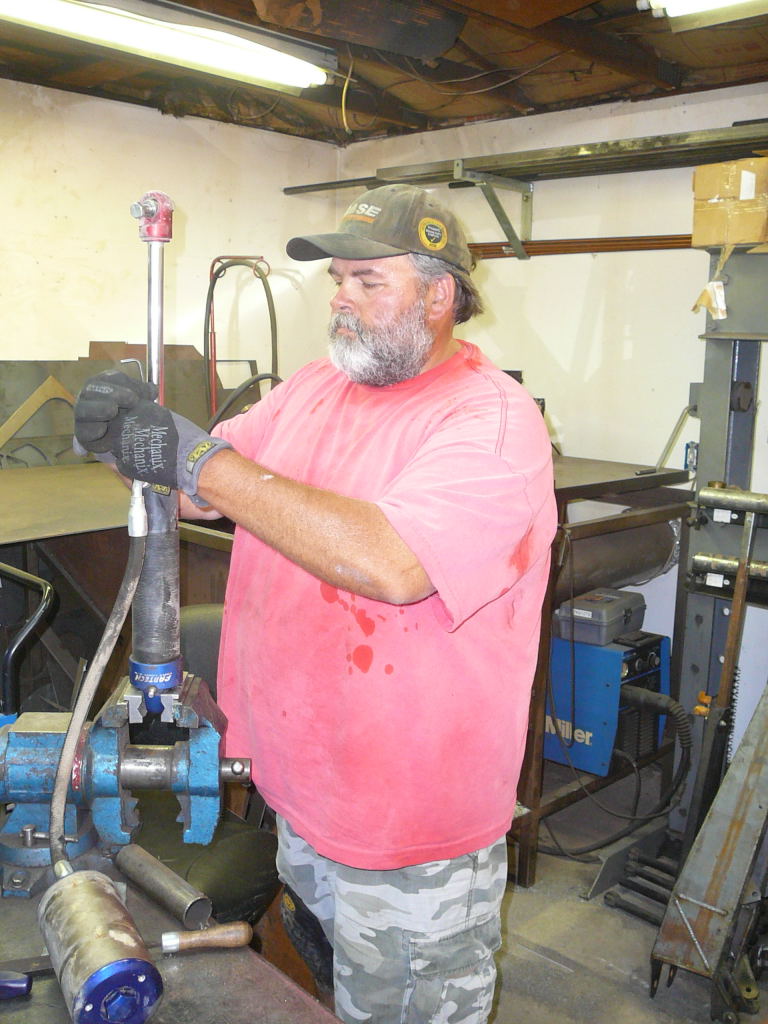 Man in a red shirt and camo shorts works on a metal part clamped in a vise in a workshop. He wears a cap and gloves