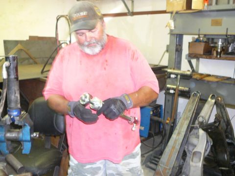 1988 Ford Bronco - A man in a workshop holds a metal linkage part, with various tools and machinery in the background.