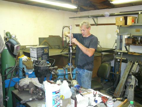 1988 Ford Bronco - Man works on a car part in a cluttered garage with welding equipment and tools visible.