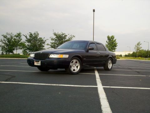 1999 Mercury Grand Marquis - A dark sedan with aftermarket wheels is parked in an empty lot under an overcast sky.