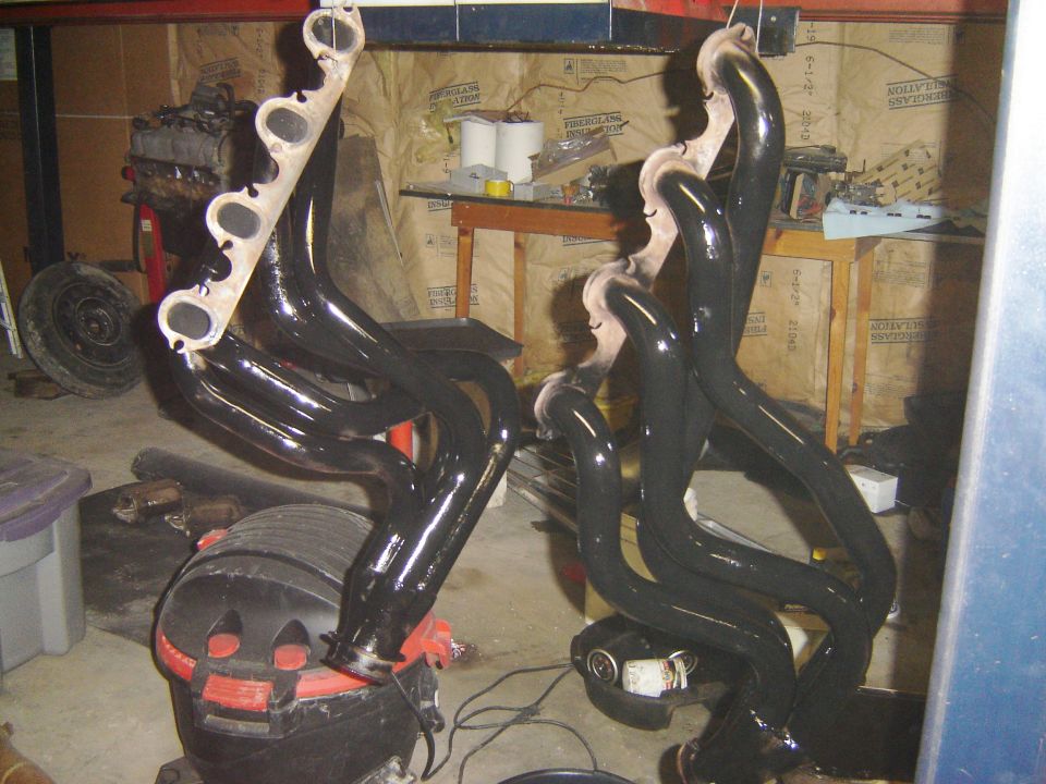 Two black exhaust headers hanging, one on a stand, the other on a red and black shop vac