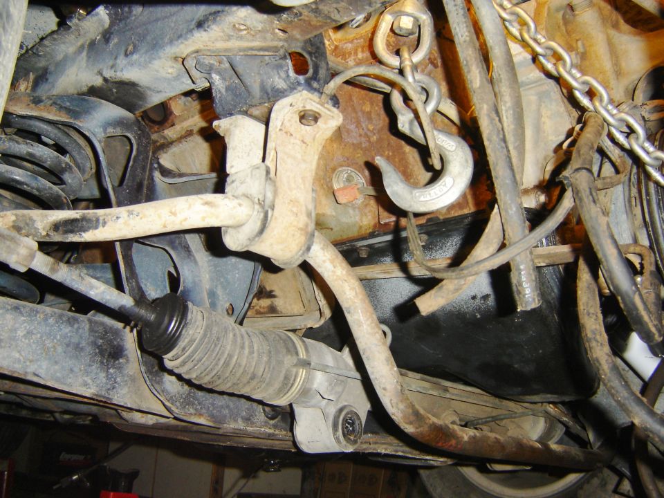 Underside view of a vehicle's steering rack and suspension components. A chain and hook are visible above