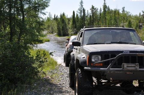 1986 Ford F-150 - A line of off-road vehicles navigates a shallow stream on a wooded trail.