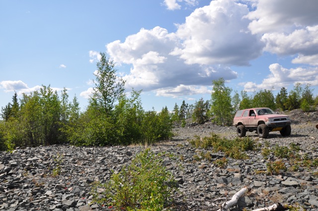Red SUV with large tires driving on a rocky, off-road path under a cloudy sky