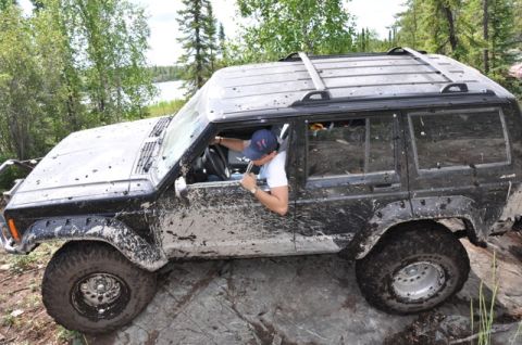 1986 Ford F-150 - A driver maneuvers a muddy Jeep Cherokee XJ over rocky terrain in a wooded area.