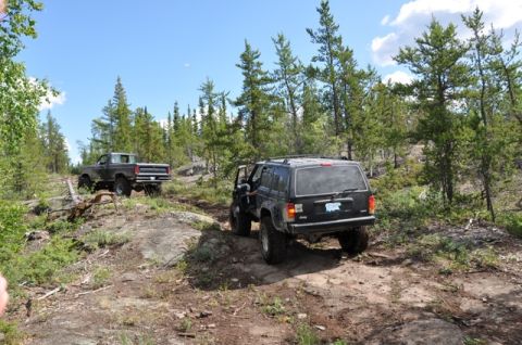 1986 Ford F-150 - Two off-road vehicles navigate a rocky, wooded trail under a partly cloudy sky.