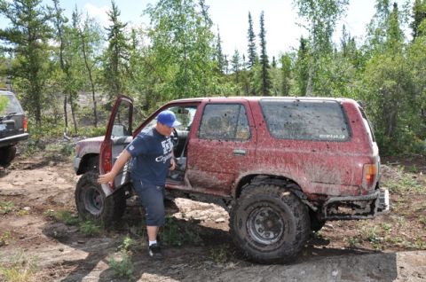 1986 Ford F-150 - A man exits a mud-covered red SUV on a rough, wooded trail, with another vehicle visible to the left.