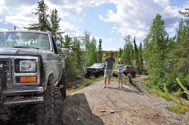 Front corner of a dirty 1980-1986 Ford F-series truck, with two people and two other off-road vehicles in the background