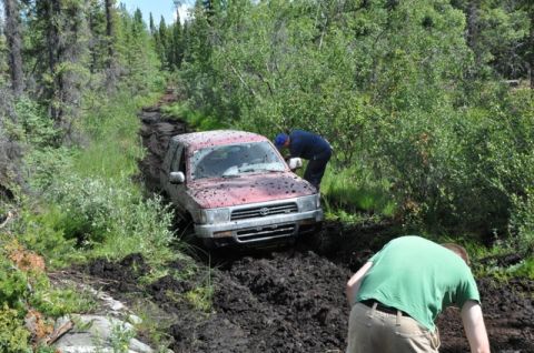 1986 Ford F-150 - A red SUV is stuck deep in mud on a wooded trail, with two individuals attempting to free it.