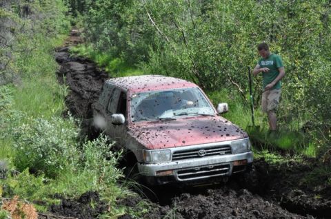 1986 Ford F-150 - A red SUV is stuck in deep mud on a wooded trail, with a person standing nearby.