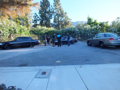 2003 Ford Crown Victoria - People gather between black and silver Panther-platform Ford sedans in a tree-lined parking lot below mountains.