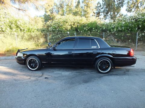 2003 Ford Crown Victoria - Side profile of a blacked-out Crown Victoria on dark wheels against greenery and a chain-link fence.