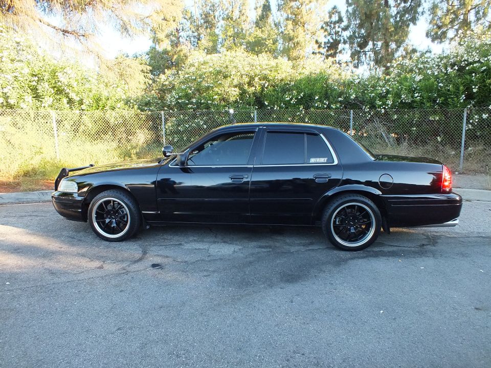 Black Crown Victoria sedan side profile on black aftermarket wheels parked beside chain-link fence
