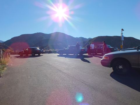 2003 Ford Crown Victoria - Silhouetted sedans and people at a roadside lot beneath sun-flared mountain peaks