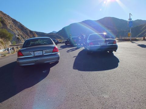 2003 Ford Crown Victoria - Two full-size sedans seen from behind facing a canyon road and guardrail
