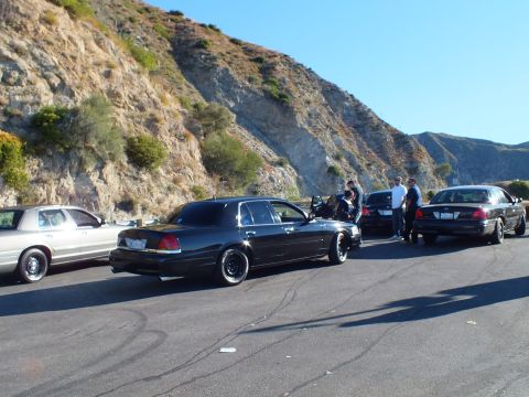 2003 Ford Crown Victoria - Lowered black sedan and silver sedan at a meet against a rugged rock slope