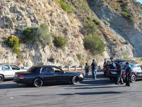 2003 Ford Crown Victoria - Lowered black full-size sedan leads a roadside meet under a rugged rock slope