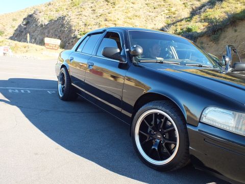 2003 Ford Crown Victoria - Glossy black sedan parked at angle with driver visible, hillside backdrop and tinted windows.