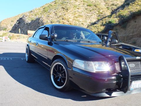 2003 Ford Crown Victoria - Dark Panther-platform sedan fitted with brush guard and headlights on, parked at hillside lot.