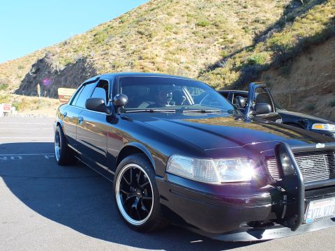 2003 Ford Crown Victoria - Front three-quarter of a dark sedan with push bar, Ford grille emblem and headlights on.