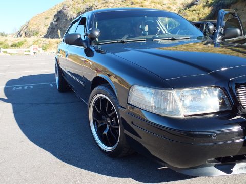 2003 Ford Crown Victoria - Glossy black full-size sedan at angle with driver inside and hillside behind at a meet.