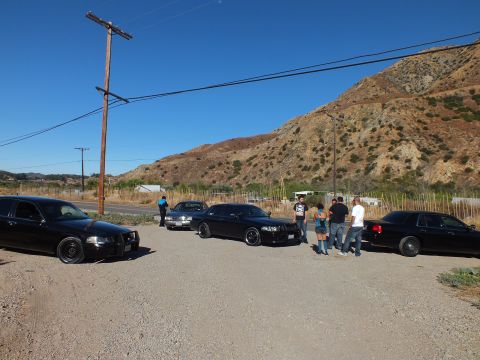 2003 Ford Crown Victoria - Several dark sedans staged on a gravel lot with people standing beside a roadside power pole.