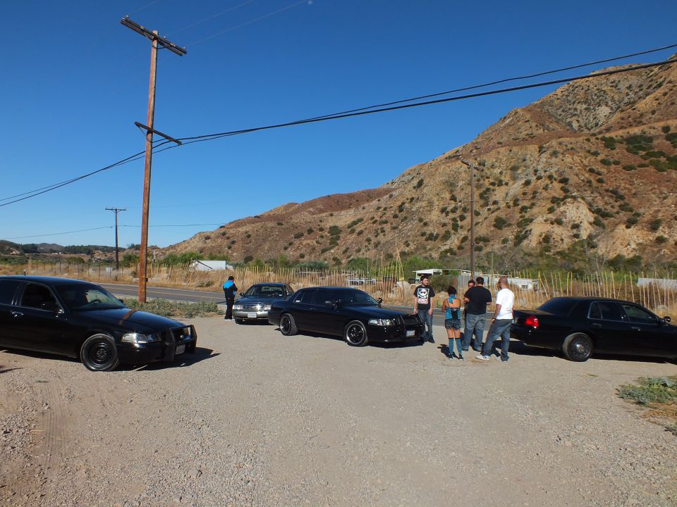 Row of black sedans parked on dirt lot near road and power pole below dry hills