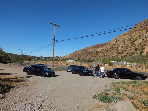 2003 Ford Crown Victoria - Dark full-size sedans parked on a dirt pullout with people gathered near a wood power pole.