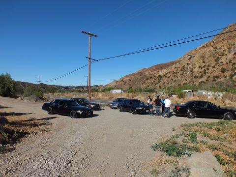 2003 Ford Crown Victoria - Several dark sedans staged on gravel near a road, people standing beneath a power pole.