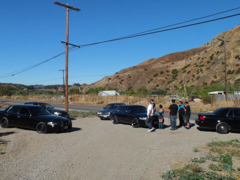 2003 Ford Crown Victoria - Dark full-size sedans angled on a dirt lot as several people gather near the roadside.
