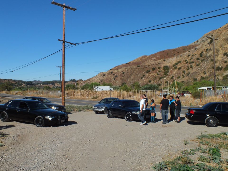 Black sedans parked on gravel lot with group of people beside road and power pole