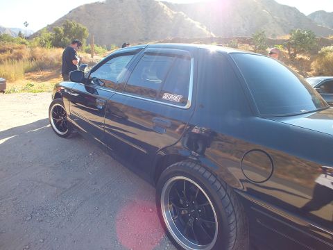2003 Ford Crown Victoria - Lowered black sedan rear quarter glows in backlight on a dirt lot, a window sticker visible.